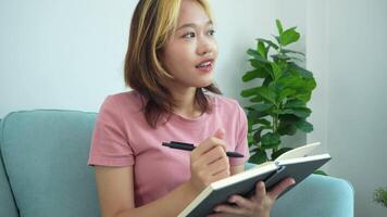 woman is writing in a notebook while sitting on a couch. She is wearing a pink shirt and has a pen in her hand. The scene suggests that she is taking notes or jotting down her thoughts video