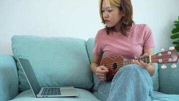 woman is sitting on a couch playing a ukulele. A laptop is on the couch next to her video