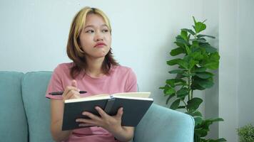 woman is writing in a notebook while sitting on a couch. She is wearing a pink shirt and has a pen in her hand. The scene suggests that she is taking notes or jotting down her thoughts video