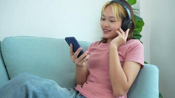 woman is sitting on a couch and looking at her phone. She is wearing headphones and she is listening to music video