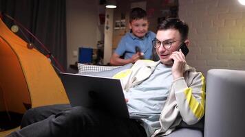 a man works at home on a laptop, the son distracts his father from work video