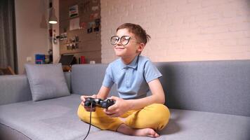 The boy with glasses is sitting on the couch playing a game on his game console video