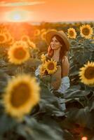 un niña en un sombrero en un hermosa campo de girasoles en contra el cielo en el noche ligero de un verano puesta de sol. rayos de sol mediante el flor campo. natural antecedentes. foto