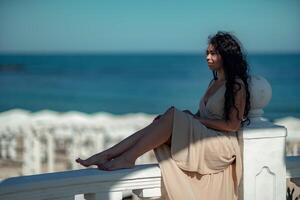 A woman in a tan dress is sitting on a white railing overlooking the ocean. The scene is serene and peaceful, with the woman enjoying the view and the calming sound of the waves. photo