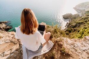 A woman is sitting on a rock overlooking the ocean with a laptop in front of her. She is enjoying the view and the peacefulness of the location. photo
