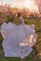 Woman blooming peach orchard. Against the backdrop of a picturesque peach orchard, a woman in a long white dress and hat enjoys a peaceful walk in the park, surrounded by the beauty of nature. photo