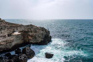 The ocean is calm and the sky is cloudy. The water is a deep blue color. The rocks are jagged and the waves are small. photo