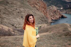 Portrait of a happy woman with long hair against a background of mountains and sea. Holding a bouquet of yellow tulips in her hands, wearing a yellow sweater photo