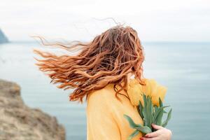 retrato de un contento mujer con pelo volador en el viento en contra el fondo de montañas y mar. participación un ramo de flores de amarillo tulipanes en su manos, vistiendo un amarillo suéter foto