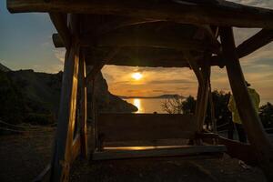 A wooden swing is suspended over a rocky shoreline. The sun is setting, casting a warm glow over the scene. photo