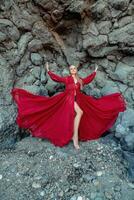 Red dress rocks woman. A blonde with flowing hair in a long flowing red dress stands near a rock of volcanic origin. Travel concept, photo session at sea