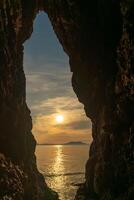 A large hole in a rock wall with the sun shining through it. The sun is setting and the sky is cloudy. photo