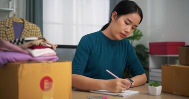 metraggio dolly sparo, giovane asiatico donna attività commerciale proprietario seduta a scrivania dai un'occhiata e Scrivi ordini i clienti in libro ,in linea shopping video