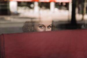 Thoughtful woman with curly hair gazing out from behind a reflective glass window, city life visible in the soft focus background photo