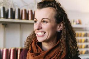 A woman with curly brown hair and a warm smile stands in a quaint craft shop adorned with shelves of colorful supplies photo