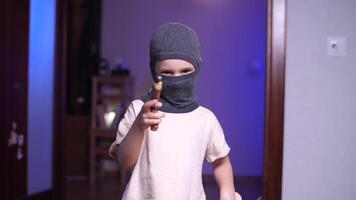 kind in een bivakmuts masker doelen een speelgoed- geweer Bij de camera, spelen bandiet video