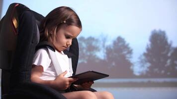 little girl sits in a child's car seat and watches cartoon videos on a tablet