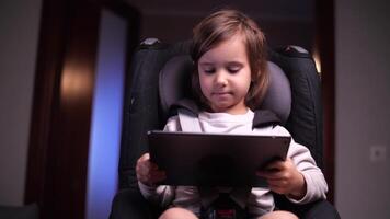 pequeño niña se sienta en un niño coche asiento y relojes dibujos animados videos en un tableta
