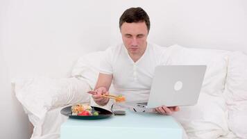 man sits in white bed in hands with laptop eats sushi from chopsticks land falls into soy sauce and splashes spot T-shirt on laptop inside computer it is bad to eat in bed you need to eat at table video