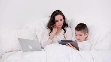 Mama und Sohn unter Decke sind Sitzung auf Sanft Bett im das Hände von Junge Tablette spielen online Freiberufler auf Laptop verbringen Zeit mit Sohn während Arbeit genesen bleibe beim Zuhause Uhr Film online Kommunikation video