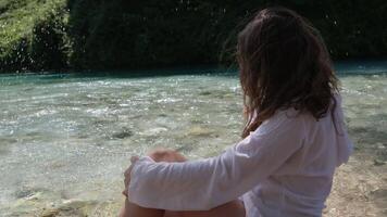 A young woman girl sits by the river in Albania, her blue eye throws splashes in all directions, she wears a white shirt, wet hair after swimming video