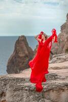 un mujer en un rojo seda vestir soportes por el océano, con montañas en el fondo, como su vestir se balancea en el brisa. foto