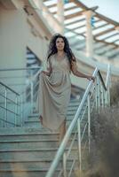 A woman in a dress stands on a set of stairs. The stairs are made of metal and are located in front of a building. The woman is wearing a necklace and a bracelet. photo