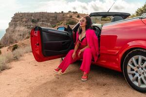 A woman in a pink suit sits in the open door of a red convertible. The scene is set on a rocky hillside overlooking the ocean. The woman is posing for a photo. photo