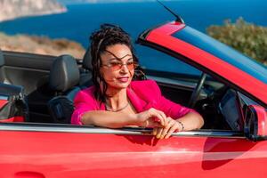 A woman in a pink jacket is sitting in a red convertible. She is wearing sunglasses and has her hand on her hip. Scene is relaxed and carefree. photo