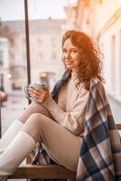 A middle-aged woman in a beige sweater with a blue mug in her hands is in a street cafe on the veranda photo