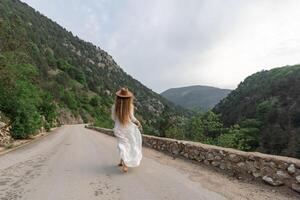 A woman in a white dress is walking down a road with a hat on. The road is surrounded by mountains and the sky is cloudy. The scene has a peaceful and serene mood. photo