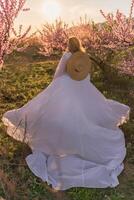 Woman blooming peach orchard. Against the backdrop of a picturesque peach orchard, a woman in a long white dress and hat enjoys a peaceful walk in the park, surrounded by the beauty of nature. photo