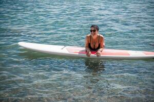 Woman sap sea. Sports girl on a surfboard in the sea on a sunny summer day. In a black bathing suit, he lies on a sap in the sea. Rest on the sea. photo