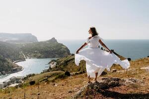 Happy woman in a white dress and hat stands on a rocky cliff above the sea, with the beautiful silhouette of hills in thick fog in the background. photo