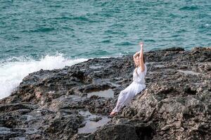 un mujer en un tormenta se sienta en un Roca en el mar. vestido en un blanco largo vestido, olas choque en contra el rocas y blanco rociar se eleva foto