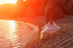 Mysterious woman silhouette long hair walks on the beach ocean water, sea nymph wind listens to the wave. Throws up a long white dress, a divine sunset. Artistic photo from the back without a face