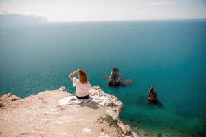 un mujer es sentado en un rock con vista a el océano. ella es señalando a el agua. el escena es pacífico y sereno, con el mujer disfrutando el ver y el calmante sonido de el ondas. foto