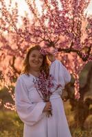 Woman blooming peach orchard. Against the backdrop of a picturesque peach orchard, a woman in a long white dress enjoys a peaceful walk in the park, surrounded by the beauty of nature. photo