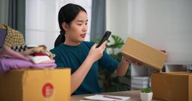 antal fot selektiv fokus skott, ung asiatisk kvinna företag ägare Sammanträde på skrivbord använda sig av smartphone skanna qr koda på lådor förpackning för frakt, uppkopplad handla video