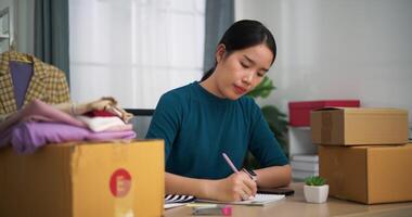 imágenes enfocar en disparo, joven asiático mujer negocio propietario sentado a escritorio cheque orden y utilizar teléfono inteligente escanear qr código en cajas embalaje para envío, en línea compras video