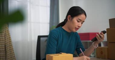 Footage dolly shot, Young asian woman business owner sitting at desk check orders by phone and write into book , Online Shopping video