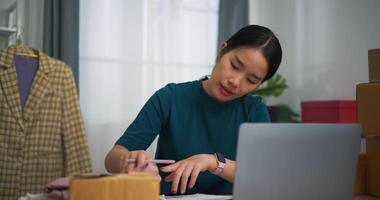 imágenes muñequita disparo, joven asiático mujer negocio propietario sentado a escritorio utilizar ordenador portátil mientras cheque pedidos y escribir dentro libro, en línea compras video