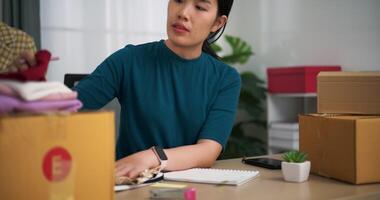 imágenes selectivo atención disparo, joven asiático mujer negocio propietario sentado a escritorio cheque y escribir pedidos clientes dentro libro ,en línea compras video
