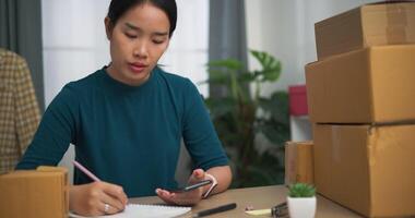 Aufnahmen Dolly Schuss, jung asiatisch Frau Geschäft Inhaber Sitzung beim Schreibtisch reden mit Kunde auf Smartphone während Verpackung zum Versand, online Einkaufen video