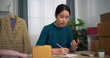 metraggio dolly sparo, giovane asiatico donna attività commerciale proprietario seduta a scrivania dai un'occhiata e Scrivi ordini i clienti in libro ,in linea shopping video