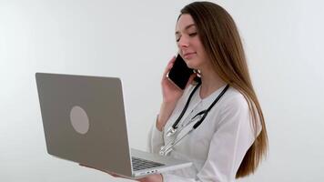 joven mestizo hembra médico en laboratorio Saco estetoscopio terminado su cuello utilizando computadora portátil, sonriente y Hablando en móvil teléfono a escritorio durante dia de trabajo en clínica video