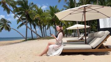 un niña participación un teléfono descansa en el playa en el Fresco aire costoso hotel viaje anuncio elegante lujo playa joven mujer niña en pareo negro traje de baño descansando mira a teléfono usos móvil redes video