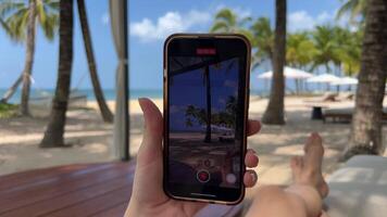 en flicka innehav en telefon vilar på de strand i de färsk luft dyr hotell resa ad chic lyx strand ung kvinna flicka i pareo svart baddräkt vilar utseende på telefon användningar mobil nät video