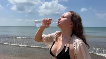 codicioso a bebida agua y derramar eso sediento costoso hotel viaje anuncio elegante mujer niña en un blanco pareo negro descansando bebidas agua mira a el teléfono usos móvil redes en el mar Oceano video