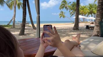un niña participación un teléfono descansa en el playa en el Fresco aire costoso hotel viaje anuncio elegante lujo playa joven mujer niña en pareo negro traje de baño descansando mira a teléfono usos móvil redes video